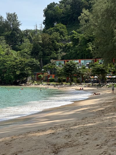 The image depicts a scenic view of a tropical beach in Ban Bang Thao, Thailand. In the foreground, the sandy shore is illuminated by the sun, casting long shadows from the surrounding trees. The ocean, with its clear turquoise water, laps gently onto the beach, creating white foamy waves. Several people are visible in the water, some swimming and others wading.
Along the shoreline, a few individuals are relaxing on the sand, with one person sunbathing on their stomach. In the mid-ground, a resort complex with modern orange buildings and white balconies is nestled amongst lush green trees. Umbrellas and lounge chairs are set up on the resort's grounds, indicating a place for relaxation and leisure. Palm trees and other tropical vegetation are abundant, framing the resort and adding to the idyllic setting. The background is dominated by a dense, verdant hillside covered in trees, suggesting a natural and unspoiled environment. The sky is clear and blue, with a few wispy clouds. The overall scene conveys a peaceful and inviting atmosphere of a tropical vacation destination.