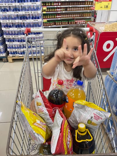 This is a vertical, eye-level shot taken inside a grocery store, likely in Novxanı, Azerbaijan, given the context. The primary subject is a young girl, possibly around 4-6 years old, peeking out from behind a shopping cart filled with groceries. She has dark hair and is smiling, looking directly at the camera with her hands held up in front of her face, as if playing a game. She is wearing a white t-shirt adorned with colorful sequins, including stars and hearts.
The shopping cart contains a variety of items, including several bags of chips, a large bottle of dark liquid (likely cooking oil), and bottles of Fanta and Coca-Cola. Some of the chip bags have Cyrillic text, indicating their origin or brand. In the background, there are shelves stocked with numerous blue and white packaged goods, some with the brand name "BIZON" visible. Further shelves to the right hold rows of canned goods. A red price tag with the text "6.90 5.99" and "TP-Araxis" is also visible. The lighting suggests it is daytime, as is typical for a grocery store. The overall scene conveys a casual, everyday shopping experience with a playful interaction from the child.