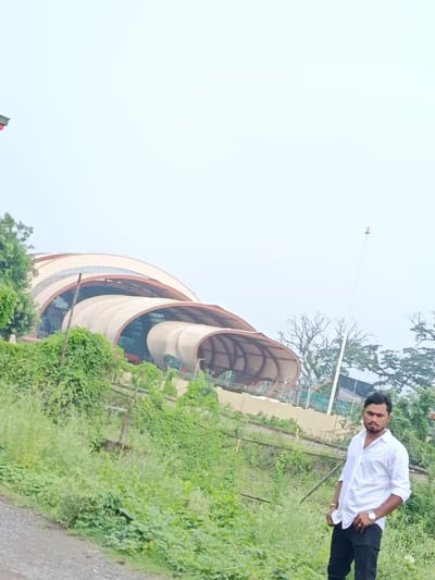 A man in a white collared shirt and black pants stands in front of a large, modern building with a curved, white roof. He is looking towards the camera with his hands in his pockets. The building appears to be an architectural landmark, possibly a museum or cultural center, with a unique, wave-like structure. Lush green foliage surrounds the area, suggesting a park or landscaped environment. The sky is overcast, creating a diffused light. A flag pole with a small flag at the top is visible in the background to the right of the building. The scene is set outdoors, with a gravel path in the foreground. The image is a candid shot, likely taken by a mobile phone.