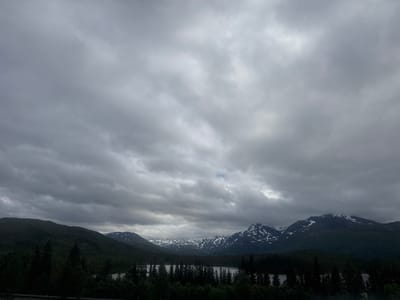 The image depicts a panoramic view of a mountainous landscape in Elsfjord, Norway. The sky is heavily overcast with dark, dramatic clouds. In the foreground, a dense forest of dark green coniferous trees lines the shore of a body of water, which appears to be a lake. Beyond the forest, rolling hills covered in vegetation lead up to majestic mountains. Some of the higher mountain peaks are capped with snow, contrasting with the dark, forested slopes. The overall atmosphere is somber and moody, characteristic of a cloudy day in a northern climate. There are no people or man-made structures clearly visible in the scene, except possibly a dark line at the very bottom which could be a road or barrier.
