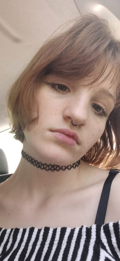 A young woman with reddish-brown hair, a nose ring, and a black choker necklace is looking downwards at the camera. She is wearing a black and white striped top and a black bra strap is visible. The background appears to be a white or light-colored wall. There is no indication in the image that it was taken in Bistrice, Montenegro.
