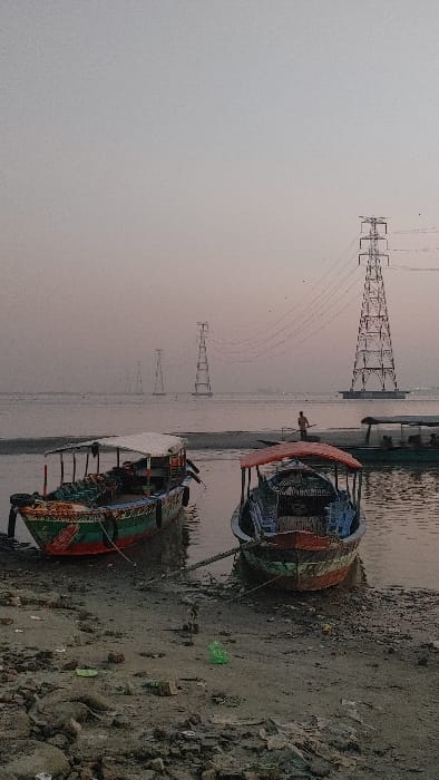 This image depicts a riverine scene in Kumarvog, Bangladesh, at twilight. In the immediate foreground, a muddy, debris-strewn shore is visible, featuring a green plastic bag and various fragments of refuse. Two traditional wooden boats are partially beached or moored. The boat on the left is larger, painted green, red, and blue with decorative accents, and features a white canopy. The boat on the right is smaller, painted red, green, and blue, equipped with a red canopy and several blue plastic chairs within its hull; it is secured to the shore by a rope. In the midground, a lone individual, silhouetted, stands in the water, appearing to pole a smaller, partially visible boat from right to left. Further in the background, a series of large metal electricity transmission pylons extends across the body of water, with power lines connecting them. The sky is hazy, displaying a gradient from light grey to a faint reddish-orange near the horizon.