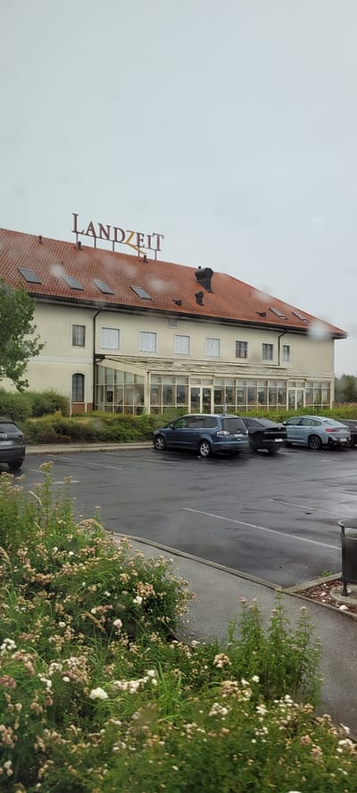 A medium shot captures the exterior of a LANDZEIT establishment in Sattledt, Austria, under an overcast sky. The building features a prominent red tiled roof with several dormer windows and skylights. A large sign with the word "LANDZEIT" is mounted on the roof. The building has a beige facade, with a glass-enclosed section on the ground floor that appears to be a dining or seating area, as evidenced by the silhouettes of people visible inside. A parking lot is in the foreground, with several cars parked. The wet asphalt of the parking lot suggests recent rain. In the immediate foreground, a variety of flowering bushes and plants are in bloom, adding a touch of natural color to the scene. The overall impression is of a functional establishment on a cloudy day.