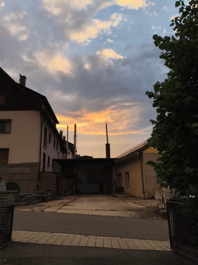This image captures a dusky scene in Pegnitz, Germany, with a dramatic sky as the backdrop. The foreground is dominated by a paved area leading into an open-sided building, possibly a workshop or storage area. Two tall, slender chimneys or exhaust pipes rise from the building's roof, silhouetted against the vibrant sky.
To the left, a multi-story building with a dark, sloped roof and cream-colored facade stands prominently. Its windows and balconies suggest residential or commercial use. A stone plaque is visible on its lower section, bearing German text that reads: "SIEMENS HAUSGERÄTE FABRIK BAYERISCHE METALLWAREN." This indicates a historical connection to Siemens household appliances and Bavarian metalware manufacturing, providing a strong location cue.
On the right side of the image, the dense foliage of a large tree partially obscures the view, adding a natural element to the otherwise industrial or urban setting. The sky is a captivating mix of colors, with soft oranges and yellows from the setting sun peeking through darker, brooding clouds. The lighting suggests it is either early morning or late evening. The overall mood is tranquil, with a hint of industrial history. The paved areas and the open building suggest ongoing or recent activity, though no people are visible. The presence of a ladder leaning against the building further implies work or maintenance.