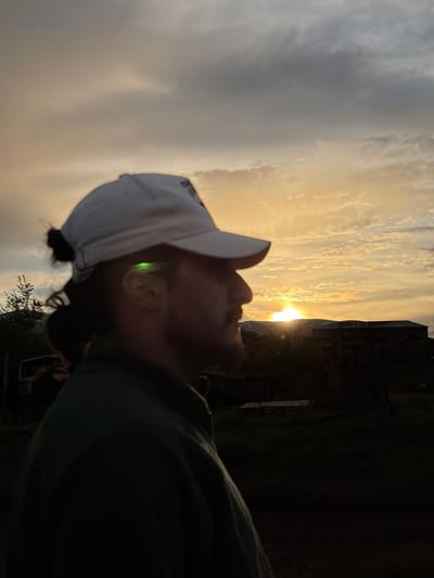 A medium shot shows a man in a green shirt and a white baseball cap looking up toward the sun. The sun is setting behind a building in the background. The photo was taken at a low angle, as if from ground level. The man is looking up, toward the sky. The photo was likely taken outdoors, during sunset. The location cannot be determined.