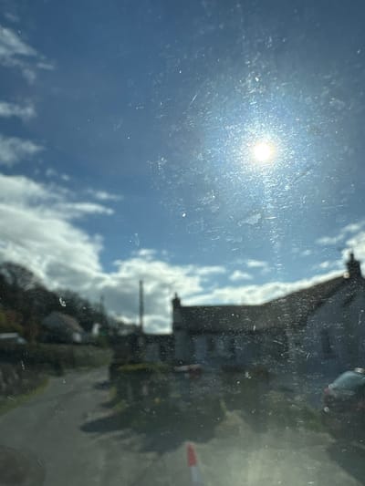 The image appears to be taken from inside a vehicle, looking outwards through a dirty or dusty window. The sun is visible in the upper left portion of the frame, obscured by the window imperfections. Reflected in the window is a scene from Snailbeach, United Kingdom, showing a building and cloudy sky. The building is dark and partially obscured. The sky is light blue with fluffy white clouds.