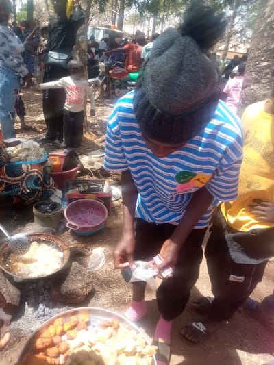 This image depicts an outdoor food preparation and sales scene in Kagoma, Nigeria.
In the foreground, a young individual, possibly male, wearing a black knitted beanie with a prominent pom-pom, a blue and white striped t-shirt featuring a "smiley face" graphic, black pants, and pink socks, stands looking down. Their hands are occupied with small, folded white papers, possibly currency or packaged items. To their immediate right, the lower torso and legs of another person wearing a yellow t-shirt and black slide sandals are visible.
To the left of the central figure, an active cooking station is visible. A dark metal pot containing light-colored food items frying in oil rests over an open fire, with visible embers and ash beneath. A slotted metal spoon is submerged in the pot. Below the central figure, a large, round metal tray displays an assortment of fried food items, including golden-brown and lighter-colored pieces. Nearby, a red plastic colander and other containers are present. Behind the cooking station, a container covered in patterned fabric holds stacked, light-colored food blocks.
The background reveals a bustling outdoor environment with numerous individuals. A man in a black shirt with a black shoulder bag stands next to a young boy in a white long-sleeved shirt. Further back, a motorcycle is parked amidst a cluster of people. The ground is bare earth, and several trees with sparse foliage are visible, indicating a sunny, outdoor setting.