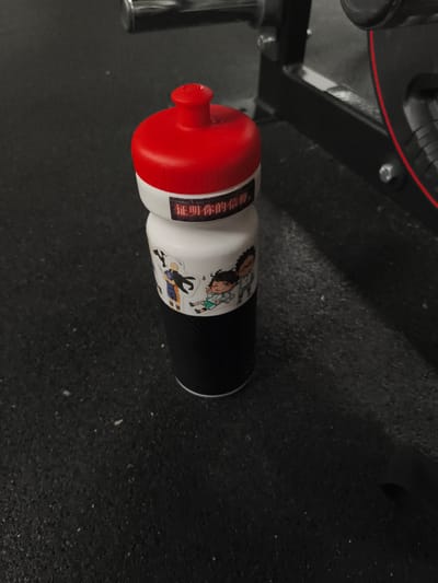 A black and white photograph features a red and white water bottle with cartoon stickers and Chinese text placed on a dark, textured gym floor. The water bottle has a bright red cap and a white body adorned with small, colorful stickers depicting cartoon characters. One sticker shows two characters embracing, while another features a single character in a green outfit, striking a playful pose. Above the stickers, a red label displays Chinese characters.
The background is dimly lit, with the glint of metal equipment visible in the upper left and right portions of the image. The setting appears to be a gym, suggested by the durable, speckled flooring and the presence of exercise machinery. The lighting is low, casting shadows and highlighting the textures of the floor and the water bottle. The overall mood is somewhat stark, with the vibrant red of the bottle's cap and label providing the only strong color contrast against the monochromatic environment. No specific time of day or weather conditions can be determined. The visible text on the bottle reads "证明你的信仰" which translates to "Prove your faith".