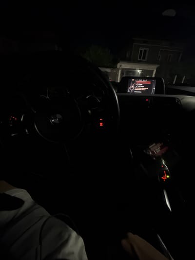 The image shows the interior of a car at night, likely in Zverino, Bulgaria, given the context. The focus is on the dashboard and steering wheel. A car's infotainment screen displays the text "iPhone," "AUX front," and "Set wireless devices via Connections menu." The steering wheel, with a BMW logo, is partially visible on the left. A person's arm and shoulder are visible in the lower left corner, wearing a light-colored shirt. The gear selector and start/stop button are illuminated in red on the center console. Outside the car, a building with windows is visible in the dark background. The scene is dimly lit, characteristic of a nighttime environment.
