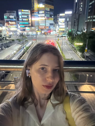 The image shows a young woman with light brown hair and fair skin taking a selfie at night. She is wearing a light-colored, textured shirt and has a yellow bag slung over her right shoulder. A light purple earbud is visible in her left ear. The background features a brightly lit urban scene with multi-story buildings adorned with numerous neon signs in Korean script. A street with traffic, including red taillights of cars, runs through the scene, and a covered pedestrian walkway is visible on the left. The general impression is of a busy urban environment at night, possibly in the commercial or entertainment district of Uijeongbu-si, South Korea.