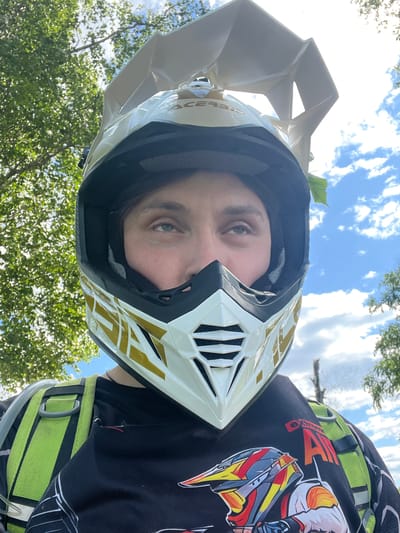 An individual is depicted outdoors, seen from the mid-chest upwards, wearing off-road gear in Новочувашский, Russia. The person is wearing a white and gold Acerbis full-face off-road helmet, with the visor angled upwards. Their eyes are visible through the helmet's opening, looking upwards and slightly to their right. A small green leaf is caught on the right side of the helmet/head area. Beneath the helmet, a black t-shirt or jersey is worn, featuring a graphic print of a helmeted rider and text including "EXTREME AIR" on the shoulder. Over the t-shirt, a neon green hydration pack or backpack with a visible drinking tube is worn. The background consists of a bright blue sky with scattered white cumulus clouds and vibrant green deciduous trees, indicating a daytime setting.