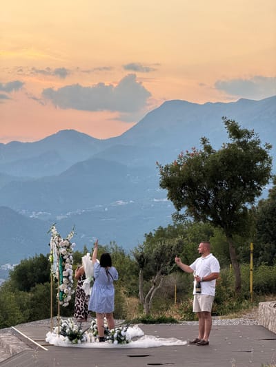 The image depicts an outdoor ceremony setup in a mountainous landscape. Two people are the primary subjects. A man in a white polo shirt and khaki shorts holds a bottle and what appears to be a glass, suggesting a toast or celebration. He is looking down at his hand. Nearby, a woman in a light blue dress is standing, with her back to the camera, also with her arm extended. She is near a floral arrangement that seems to be part of an arch or decorative structure. Another person in a leopard-print dress is partially visible behind her.

The scene is set against a backdrop of rolling hills and mountains, with a hazy sky that suggests either early morning or late afternoon light. The warm, soft tones in the sky indicate a time near sunrise or sunset. The presence of lush green trees and the mountainous terrain point to a natural, scenic location. The ground appears to be paved, with some loose gravel and scattered white petals. The overall atmosphere seems celebratory, possibly a wedding or a special event. No text is visible in the image.