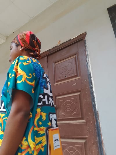 A medium shot depicts a dark-skinned woman standing outdoors, wearing a colorful headscarf and a blue garment with a Chanel logo and perfume bottle design. She appears to be in Ogboko, Nigeria, evidenced by local architectural details. Behind her is a weathered brown door with decorative molding and aging paint. The wall adjacent to the door is light colored. The scene is captured in natural daylight.