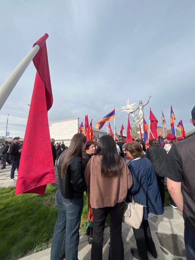 A large crowd gathers near a statue of Mother Armenia in Akhuryan, Armenia. The monument is surrounded by numerous red and orange-blue-red flags, symbols of Armenian identity. The crowd is composed of individuals dressed in casual clothing, and their activities or interactions with one another are not clearly defined. The environment is partly visible with a sky overhead and grassy areas and sidewalks alongside the group.