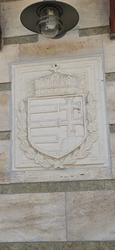 The image shows a relief carving of the Hungarian coat of arms on a stone wall. The carving is detailed, featuring a crown, a shield with horizontal stripes and a double cross, and a wreath of leaves and flowers. Above the relief, a dark metal outdoor light fixture with a glass bulb is visible, mounted on a stone wall. The wall is constructed from large, rectangular stone blocks, with bands of darker stone interspersed with lighter stone. The style of the relief and the architectural elements suggest a historical building, possibly in a public space. The scene is set outdoors during the daytime, with natural light illuminating the stone and the carving. Based on the information provided, this relief is located in the city of Cserszegtomaj, Hungary. There are no people or animals present in the image.