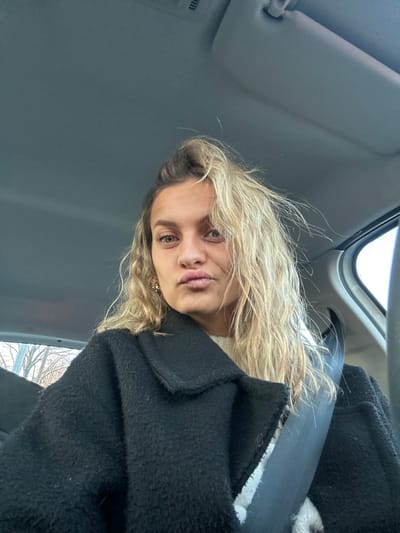 A woman with wavy blonde hair and brown eyes sits in a car. She is wearing a black coat and a white sweater. The seatbelt is visible across her chest. The interior of the car is gray. She has a slight pout. The view from the car window includes trees. She is turned slightly to the right. The sun visor is visible above her head.