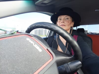 A medium shot captures a woman wearing a black fedora and a gold chain, driving a vehicle. She is looking forward and slightly to the right, with a serious expression. The interior of the car is visible, including the steering wheel, dashboard, and a portion of the passenger seat. Outside the car, apartment buildings and power lines can be seen, suggesting an urban or suburban environment. The context provided indicates the location is Zanevka, Russia.