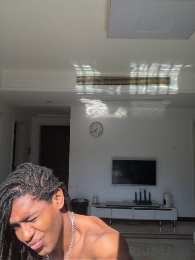 A vertical interior shot shows a person with dark dreadlocks and a silver chain lying down in a modern apartment. The walls are painted white, with sunlight streaming in through a vent near the top of the frame. A television, a wall-mounted white cabinet, and an analog clock are visible on the wall. The photo appears to be taken indoors, but the specific location in Binjiang District, China cannot be determined from the image.