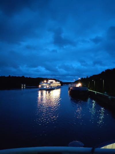 A wide-angle, low-light, vertical photograph captures two illuminated boats on a dark, calm body of water at dusk in Devyatiny, Russia. The sky is a deep, moody blue with scattered, darker clouds. On the right side of the frame, a dark cargo barge is docked along a concrete embankment, with several streetlights visible above it. On the left side of the frame, a larger, multi-decked passenger boat is moving forward. This boat is brightly lit with numerous windows casting a warm, yellow glow onto the water's surface, creating a shimmering reflection. The dark shoreline with trees forms a silhouette against the sky on both sides of the water. A railing is visible in the foreground, suggesting the vantage point is from a bridge or higher structure.