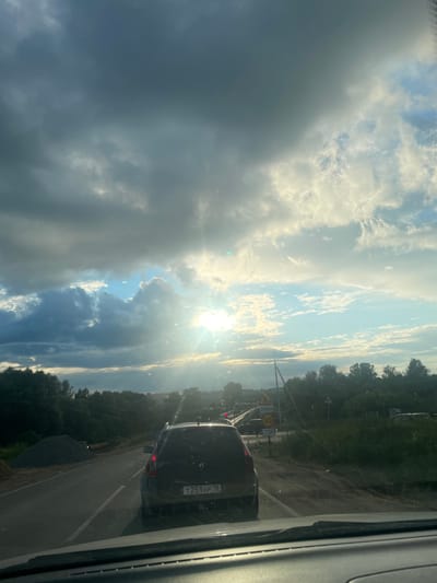 The image depicts a view from inside a vehicle, looking out at a road and a dramatic sky. The scene is set in Гавриловка, Russia, and it appears to be late afternoon or early evening due to the low angle of the sun. The sky is filled with a mixture of dark, heavy clouds and bright, sunlit patches, suggesting a dynamic weather pattern, possibly the end of a storm or approaching twilight. The sun's rays are breaking through the clouds, creating a lens flare and illuminating the scene.

On the right side of the frame, a dark-colored SUV is visible in traffic on a paved road. The vehicle has a license plate that reads "T251HP 18". There are road signs visible, including a yellow warning sign with a triangle and a zigzag line, and a circular sign with "30" inside, indicating a speed limit. Another sign reads "СТОП," which translates to "STOP." The road is bordered by lush green trees on the right and a somewhat unkempt embankment on the left, with piles of gravel.

The overall mood is somewhat somber due to the heavy clouds, but also hopeful and bright due to the sunlight breaking through. The image captures a moment of travel on a rural or suburban road, under a striking sky, in what appears to be a transitional time of day.