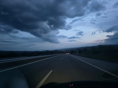 The image presents a first-person view from inside a vehicle traveling on a multi-lane highway at dusk or twilight. The dominant feature is the dramatic, heavily overcast sky, filled with dark, dense clouds in shades of deep gray and blue. Patches of lighter blue are visible where the cloud cover is less thick, and a faint reddish-pink glow can be seen near the horizon on the right, suggesting the last remnants of a sunset.
The asphalt highway stretches into the distance, clearly marked by white dashed and solid lines, and appears somewhat reflective due to the low light. A metal guardrail runs along the left side of the road. In the mid-ground, red taillights from several distant vehicles are visible on the highway, indicating other traffic. The landscape surrounding the road consists of dark, undulating hills and mountains, which appear silhouetted against the dimming sky, suggesting a rural or mountainous setting.
The foreground shows the top portion of the car's dashboard and windshield, with a soft glow from the vehicle's headlights illuminating the immediate road ahead. No people are visible. There is no discernible text within the image. The scene captures a journey through a quiet landscape under a moody, darkening sky, consistent with the Blatino, Bulgaria location context.