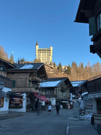 The image depicts a street view in Gstaad, Switzerland, looking uphill towards the Gstaad Palace Hotel under a clear, bright blue sky. A crescent moon is visible in the upper left quadrant of the sky, above a bare tree branch.

The Gstaad Palace Hotel, a multi-story, light-colored building with two prominent corner towers and a central structure, is situated on a wooded hill in the background. The hotel exhibits numerous windows arranged symmetrically.

In the mid-ground and foreground, traditional Swiss chalets constructed primarily of dark wood with pitched roofs line a paved street or plaza. Several rooftops have visible patches of snow. On the left side, a chalet houses a ground-floor shop with a red awning and a sign displaying "TINGRAF". A street lamp stands in the lower left. On the right, parts of other wooden chalets are visible, including one with dark wood and open green shutters on its upper story, framing the right edge of the image.

Two individuals are walking away from the camera in the center of the paved area. One wears a white jacket and dark lower garments, while the other wears dark clothing. A third, indistinct figure is partially visible under the red awning on the left. The paved surface shows faint traces of what appears to be ice or compacted snow. Bare deciduous trees and evergreen conifers are interspersed throughout the scene and on the hills.