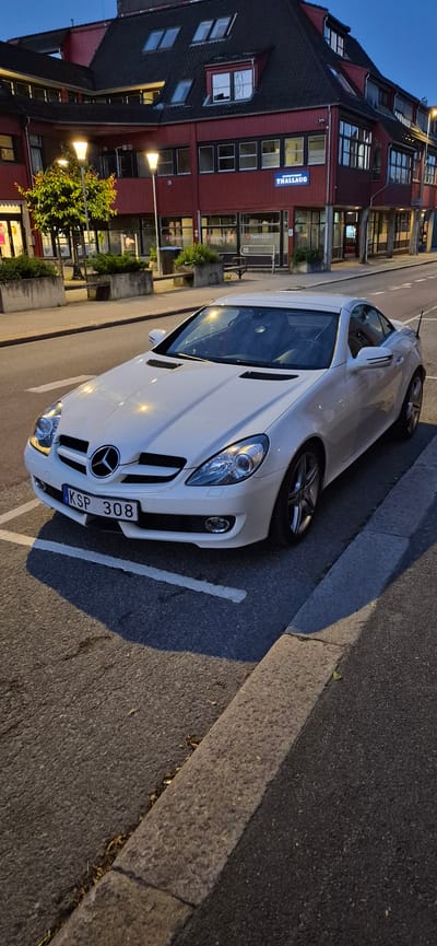 A white Mercedes-Benz SLK-Class convertible, license plate KSP 308, is parked on a street in front of a red building with commercial establishments. The building features large windows and signage that reads "THALLAUG" in blue illuminated letters. One storefront visible is "Fretexjobb." The scene appears to be in Lillehammer, Norway, indicated by the presence of the building and streetscape. The time of day is late evening, as suggested by the artificial lighting and the twilight sky. The car is parked at an angle, with its front facing the viewer. The street is paved, and a concrete curb separates the road from a sidewalk. There are streetlights visible, and some trees and landscaping are present in front of the building. No people are visible in the image.