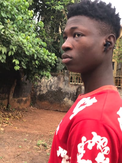 An outdoor portrait, taken in Abatete, Nigeria, shows a young person, likely male, with dark skin and short, dark, textured hair. The subject is looking directly at the camera with a neutral expression, their head tilted slightly to their left. They are wearing a vibrant red short-sleeved shirt patterned with abstract white designs, which are visible across the shoulder and upper arm. A black earbud is noticeable in their left ear.
The background features lush green foliage, including bushes and parts of trees, on the left side of the frame. Towards the center and right, a weathered, dark-colored concrete or stucco wall with visible texture and discoloration stands, behind which a metal grate or window frame of a building can be vaguely seen. The ground in the foreground and to the right is a reddish-brown dirt path partially covered with dry leaves. The scene is illuminated by natural daylight, suggesting an overcast sky or indirect sunlight, as there are no harsh shadows. No decipherable text is visible, only the decorative patterns on the shirt.