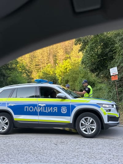 A police car, marked with "ПОЛИЦИЯ" in Cyrillic script, is parked on the side of a road. Two police officers, one wearing a high-visibility vest, are interacting near the driver's side of the vehicle. The scene appears to be in a rural or semi-rural area, with dense green foliage visible behind the car. Sunlight filters through the trees, suggesting it is daytime, possibly late afternoon. Two signs are visible on a pole in the background. One sign has text that is partially legible, and the other is orange with a symbol that might indicate a restriction or warning. The overall atmosphere seems to be one of an official police stop or operation.