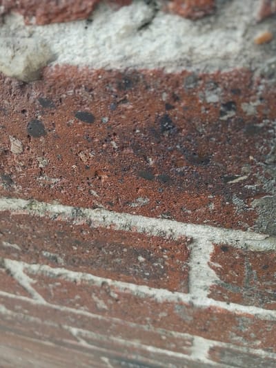 The image shows a close-up, angled view of a brick wall. The bricks are predominantly reddish-brown, with visible mortar lines that are lighter in color and appear somewhat rough and textured. Some of the bricks have dark inclusions or imperfections, giving them a mottled appearance. The lighting suggests it is daytime. The perspective is from a low angle, looking upwards at the wall. The image does not contain any people, animals, or discernible actions. The environment appears to be an exterior wall, likely part of a building. There is no specific information visible that would allow for precise identification of its location within Rumuepirikom, Nigeria.