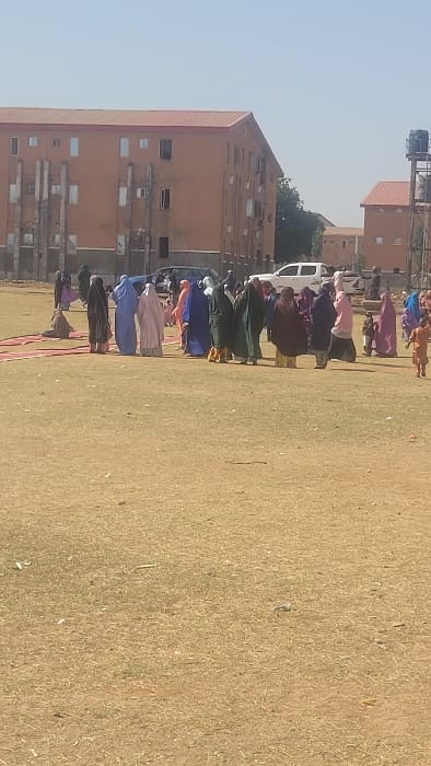This image depicts an outdoor gathering on a dry, grassy field in Badiko, Nigeria. In the foreground and midground, the ground is covered with sparse, yellowish-brown grass and dry earth. Approximately 20-30 individuals, predominantly adults and several children, are present. Most adults, particularly women, are dressed in long, flowing garments and head coverings of various colors including blue, pink, brown, green, and purple.
In the mid-left, several red fabric strips or carpets are laid out on the ground, forming a path or designated area. A person in a brown garment is observed sitting on the ground nearby. The main group of people is clustered in the midground, with individuals standing in small groups, appearing to converse or observe their surroundings. A young child in a light brown outfit is visible walking towards the right side of the frame. Further back, another person in dark attire walks near a small pile of objects on the ground.
In the background, two multi-story buildings with terracotta-colored exteriors and red roofs are visible under a clear blue sky. The prominent building on the left features multiple windows and vertical concrete structural elements on its facade. To its right, a dark-colored sedan and a white pickup truck are parked. Further right, a smaller building with a similar aesthetic is partially visible behind a metal water tower structure.