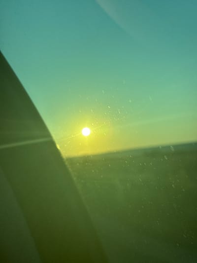 This media file captures an abstract view through a window, likely a car windshield, on a sunny day in Lativka, Ukraine. The primary focus is the interplay of light and shadow on a surface that appears slightly textured, possibly due to dust or water droplets. The scene is dominated by a gradient of colors, transitioning from a cool teal on the left to a warmer yellow and then to a muted olive green on the right.
A prominent, bright, circular light source, likely the sun, is visible in the center of the frame, casting a soft glow and a thin beam of light downwards. This light source creates a distinct highlight. A diagonal shadow line cuts across the frame from the upper right to the lower left, dividing the teal and yellow-green areas. This shadow suggests an obstruction outside the window, possibly a building or tree. The surface also displays numerous small, light-colored speckles scattered across it, which could be dust particles or imperfections on the glass.
The time of day appears to be either morning or late afternoon, given the angle and intensity of the sunlight. The weather is clearly sunny, and the overall mood is somewhat serene and contemplative, evoked by the soft light and muted colors. There are no visible people, objects, or text in the frame. The scene offers a minimalist, abstract depiction of a common visual experience, highlighting the beauty of light, color, and shadow.