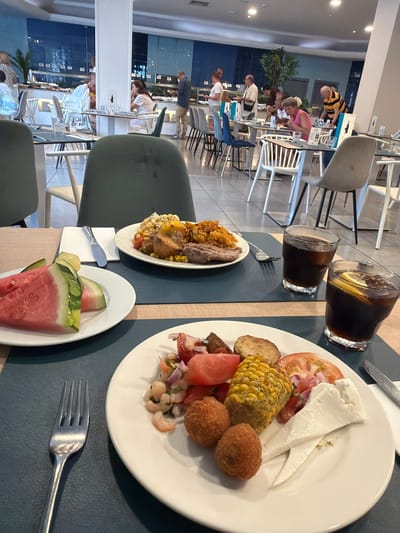 The image depicts a buffet restaurant scene, likely in Las Dehesas, Spain, with diners selecting food and seated at tables. In the foreground, two white plates are laden with food. One plate contains sliced watermelon and what appears to be a melon. The other plate features a variety of items including what looks like falafel, corn on the cob, a slice of tomato, some form of salad with shrimp, and cheese. Two glasses of a dark beverage with ice and lemon slices are also present on a dark placemat. In the background, other diners are visible, some standing at a buffet counter and others seated at tables. The environment suggests a casual dining setting with light-colored tables and chairs, and a blue-tiled wall in the background. The lighting appears to be overhead, creating a well-lit space.