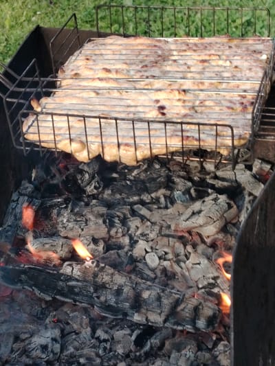A close-up shot shows a grill with food being cooked over hot coals. The food appears to be a large, flat piece, possibly pizza or flatbread, resting on a metal grill grate. The coals below are glowing red and orange, with some visible flames licking upwards. The grill is situated in an outdoor setting, with a blurry green grassy area in the background. The image is taken from a slightly elevated perspective, looking down into the grill. There are no people visible in the image. The overall scene depicts an outdoor cooking activity.