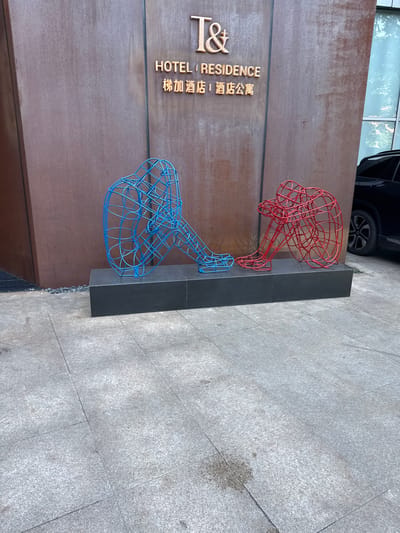 The image shows two wireframe sculptures of human figures, one in blue and one in red, seated facing away from each other on a dark rectangular plinth. The sculptures are positioned on a paved outdoor area with gray stone tiles. Behind the sculptures, a building facade with a textured, rusty-brown surface is visible. Above the sculptures, a sign reads "T& HOTEL | RESIDENCE" in gold lettering, with Chinese characters below it. To the right of the building facade, a portion of a dark-colored vehicle is parked. The scene appears to be in an urban environment, likely in the Minhang District of China.