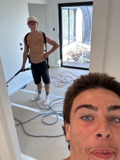 Two men in an unfinished room. One man, closer to the camera, is shirtless and wearing shorts. He is holding a tube, wearing a baseball cap, and has a harness on his back. The other man is closer and in focus. He is taking the photo. The room has a large window showing an outdoor scene with building material. There are cords on the floor. The room appears to be in the final stages of construction.