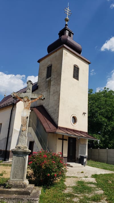 This media file captures a sunny day in Vyšné Nemecké, Slovakia. The scene is dominated by a church tower with a dark, multi-layered roof and a golden cross adorning its peak. The tower is painted a light cream color and features several windows, some with horizontal louvers and a small circular one.
In the foreground, a stone crucifix stands weathered and aged, depicting Jesus. The statue appears to be painted, though the colors have faded over time. Beside the crucifix, a vibrant bush of red roses adds a splash of color to the scene.
Behind the church, a tall green tree provides a backdrop, its leaves lush and full under the clear blue sky dotted with fluffy white clouds. A paved walkway leads towards the entrance of the church, where a dark wooden door is visible. There is some visible text on the base of the crucifix, but it is illegible due to the angle and resolution. The overall impression is one of a peaceful, rural setting bathed in bright sunlight.