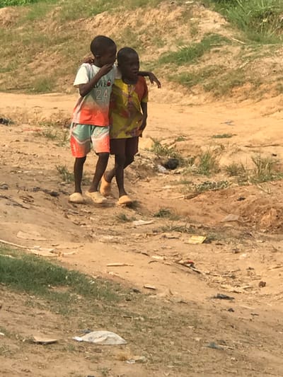 Two young children are walking side-by-side on a dirt path. The child closer to the viewer wears a white shirt with a graphic, red and teal patterned shorts, and sandals. The other child is wearing a yellow patterned shirt. The path has grass growing on the edges and scattered debris. The overall tone of the image suggests a rural, potentially impoverished setting.