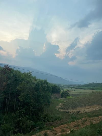 The image displays a vast rural landscape under a dynamic, cloudy sky, likely taken in Thôn 18, Vietnam. The sky dominates the upper portion, featuring light blue patches interspersed with large, soft white and grey cumulus clouds. Distinct sun rays pierce through the clouds on the left side, creating ethereal light shafts.

In the middle ground, a long, dark blue-grey mountain range stretches horizontally across the frame, appearing hazy due to atmospheric perspective. Below the mountains, a dense band of dark green trees, possibly a forest, runs across the mid-section, partially obscuring the lower slopes.

To the right, the terrain consists of gently sloping or terraced land, characterized by a mix of vibrant green vegetation and exposed reddish-brown soil, suggesting agricultural use or recent excavation. Further back on the right, additional green hills or fields are visible.

Several thin, dark power lines or communication cables traverse the scene horizontally, running above the treeline and across the distant landscape. A few slender, light-colored pylons or utility poles can be faintly discerned in the far upper right background. No people or specific activities are depicted; the scene is a tranquil, static landscape. The diffused light and visible sun rays suggest either early morning or late afternoon. No text is visible in the image.