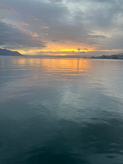 The image displays a panoramic view of Lake Geneva at either sunrise or sunset, observed from the water near Montreux, Switzerland. The foreground consists of the lake's calm, dark blue-green surface, exhibiting minimal ripples. A bright, horizontal band of intense orange and yellow light, indicative of a low sun, extends across the distant horizon, casting a distinct vertical reflection across the water towards the viewer. To the far left, a dark, elongated mountain range emerges from the lake's edge. To the far right, the shoreline features a collection of distant structures, identifiable as urban buildings, along the water. The sky is largely overcast with varying shades of grey and dark blue clouds, with pockets of brighter, orange-hued clouds near the light source on the horizon. No individuals, explicit actions, or specific interactions are discernible within the scene.