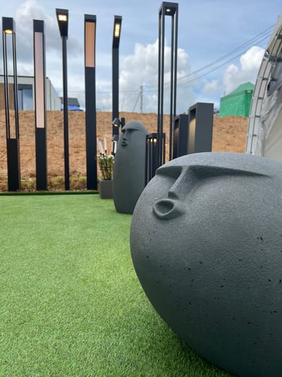 The image depicts an outdoor scene featuring a collection of modern art sculptures and lighting fixtures arranged on a patch of artificial turf. In the foreground, a large, dark gray, egg-shaped sculpture with a stylized face is prominently displayed. To the left and slightly behind it, another similar sculpture is visible. In the middle ground, a row of tall, black, rectangular light posts with illuminated panels stand against a backdrop of reddish-brown soil and some green foliage. Further back, a green tent or structure is visible. The sky above is partly cloudy. The location is identified as Yakunino, Russia. There are no people visible in the image.