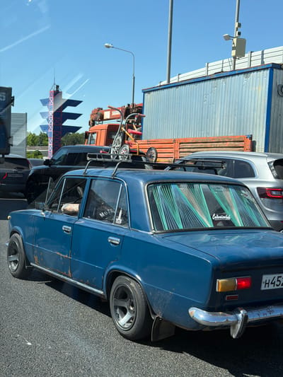 This image depicts a daytime street scene in Десна, Russia, under a clear blue sky. The primary subject is an aged, blue VAZ-2101 sedan (Lada) with significant rust and paint wear visible along its lower body panels. It is equipped with dark gray aftermarket alloy wheels and a black roof rack on which a child's bicycle and another small wheeled item are secured. The front passenger window is open, revealing a tattooed forearm. Through the closed rear passenger window, an occupant is visible wearing a shirt with "MOTHER RUSSIA" and a "CCCP" (USSR) space logo. The rear window features vertical green and white string decorations and a sticker with the Cyrillic word "Советский" (Soviet). The vehicle's rear license plate partially displays "H452".
In the midground, to the right and slightly behind the blue sedan, a silver SUV is present. Further back, a black SUV with a partially visible license plate "T764PY 199" is observed. To the right of the black SUV, an orange flatbed truck, featuring a crane marked "FISKARS," carries a small bicycle or scooter on its bed.
In the background, a distinctive blue and red tower-like structure with horizontally stacked fin-like elements displays the Cyrillic word "ЦЕНТР" (Center). Adjacent to this structure, a corrugated metal building with a blue frame supports a pole bearing a streetlight and a traffic camera system. Green foliage is visible below the tower structure. All vehicles appear to be in traffic on an asphalt road.