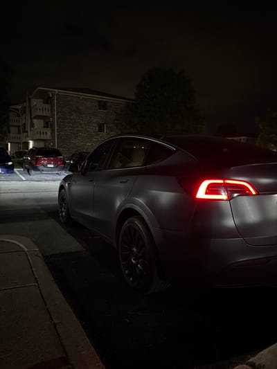 A matte gray Tesla Model Y is parked at night in a residential area in Vernon Hills, Illinois. The car's rear taillights are illuminated, casting a red glow. The vehicle is parked in a lot next to a curb and asphalt pavement. In the background, an apartment building with balconies is visible, along with other parked cars, including a red SUV and a blue sedan. Trees and foliage are also present, partially obscuring the building and sky. The scene is dimly lit, characteristic of nighttime conditions.