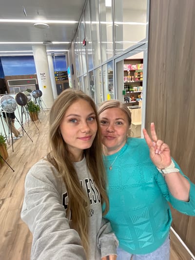 In this selfie, two women are posing indoors. The woman on the left has long blonde hair and is wearing a grey sweatshirt. The woman on the right has blonde hair and is wearing a teal sweater, making a peace sign with her right hand. They are standing in a hallway with a wooden floor and wooden paneling on the right. In the background, there are displays of what appear to be globes on tripods and plants. There is also a shop visible through a glass partition, stocked with various items on shelves. The overall environment suggests an indoor public space, possibly an airport or a shopping area. There is no specific indication that the location is in the city of Старое Мартьяново, Russia.