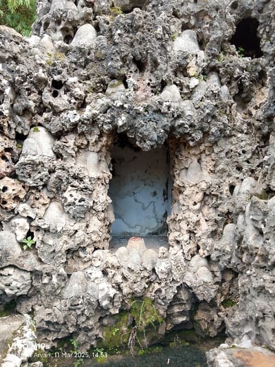 The image shows a close-up of a rocky wall with a small opening, located in Cirebon, Indonesia, at the coordinates -6.736 latitude and 108.544 longitude. The wall is composed of grey and beige, porous rock, possibly limestone, with various indentations and formations. Some green plants are visible at the bottom of the image, along with a small pool of water. The opening in the wall reveals a white concrete or plastered surface with a small ledge on which a red object rests. The image is taken at a close distance to the wall and appears to be a still shot taken during the day. The image is well-lit, but not overly bright. There is a small watermark in the bottom left corner of the image.