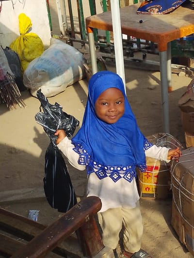 The image captures a young child standing outdoors in Unguwa Uku Kauyen Alu, Nigeria. The child, appearing to be female with dark skin and a smiling expression, is centrally positioned and faces the viewer. She wears a bright blue hijab featuring a white band with blue and white geometric embroidery at the lower edge, a long-sleeved light-colored top, light-colored trousers, and dark open-toed sandals. Her right hand holds a crumpled black plastic bag.
In the background, to the left, a bundle of sticks and two sacks—one yellow and tied, the other large and light-colored—rest against a light-colored wall. Further back, a green metal fence is visible. An orange-topped plastic table with a folded blue and white patterned fabric and a small dark object sits in the mid-background. To the right of the child, several cardboard boxes are stacked, some secured with white string or netting. The ground consists of light brown, dusty earth. A wooden structure is partially visible in the lower-left foreground. The scene is illuminated by clear daylight.