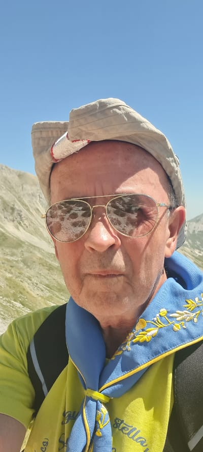 This is a selfie taken outdoors with a clear blue sky above. The subject is a man wearing a light-colored cap, aviator-style sunglasses, a bright yellow t-shirt, and a blue neckerchief with yellow embroidery. The background shows a mountainous landscape, typical of the Gran Sasso mountain range near L'Aquila, Italy, characterized by rocky slopes and sparse vegetation. The man appears to be in good health and is looking directly at the camera. The overall impression is of a person enjoying an outdoor activity in a scenic, elevated location.
