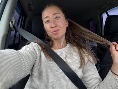 The image features a single female individual seated in the passenger side of a vehicle, appearing to take a selfie. She is positioned facing forward and slightly to her right, looking directly at the camera. Her hair is long, light brown, and styled loosely, with some strands pulled forward over her left shoulder, and a section held by her right hand near her chin. She has light-colored eyes, a light complexion, and visible freckles across her nose and cheeks. Her lips are closed in a subtle smile. A small, gold-colored hoop earring is visible on her right earlobe.
She is wearing a light, possibly off-white or light gray, ribbed knit sweater. A black seatbelt strap crosses diagonally over her chest and left shoulder, securing into a mechanism on her left side. The seatbelt's height adjuster is visible on the vehicle's A-pillar to her left. On her left wrist, a thin black string bracelet with a small metallic bead is visible.
The interior of the vehicle appears dark, with black upholstery visible on the seats behind her and to her right. The overhead console and a grab handle are partially visible in the upper right background. Through the side windows, an overcast or low-light exterior is suggested by muted colors and soft light, with no distinct landscape features. A faint purple object is discernible between the front and rear seats on the far right side of the frame.
The image provides no external visual cues or contextual information to identify the specific location as Karagay, Russia.