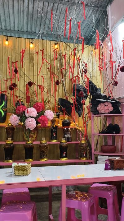 The image shows a decorated area, possibly a shop or restaurant, in Đưng K'Si, Vietnam. A table with a white top and pink legs is set up, with pink stools around it. On the table, there's a small woven basket. The background features a wall with vertical wooden panels and overhead lighting. A tree-like structure with bare branches is adorned with numerous red ribbons and some red decorative balls. Several shelves are visible, holding various glass jars with amber-colored contents and gold lids, as well as some bottles. A bouquet of pink and red artificial flowers is placed in a vase on one of the shelves. There are also some black fabric decorations and a bundle of pink artificial flowers wrapped in black paper on a higher shelf.