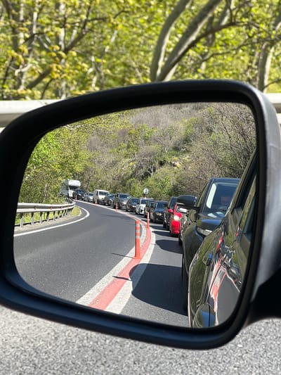 The image depicts a traffic jam reflected in a vehicle's side mirror, likely on a highway near Krupnik, Bulgaria, given the presence of dense foliage on the roadside. The traffic is congested, with numerous cars lined up behind each other, and an orange traffic cone is visible on the road. The road appears to be undergoing construction or maintenance, suggested by the orange cone and lane markings. The surrounding environment includes green trees and vegetation. The mirror itself has a black frame.